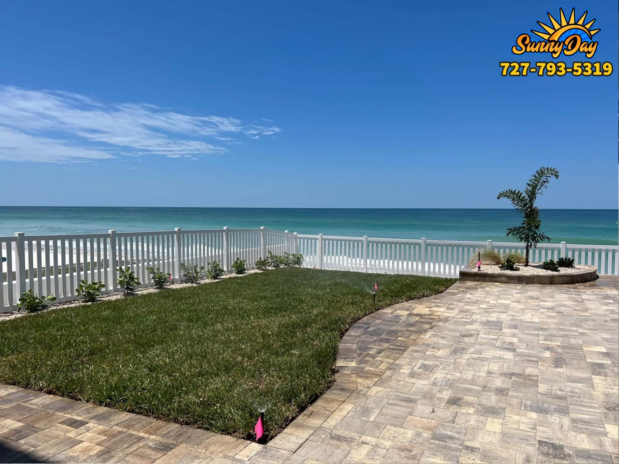 Beachfront hardscape landscaping with paver patio, white fencing, sod, irrigation flags, and Gulf view
